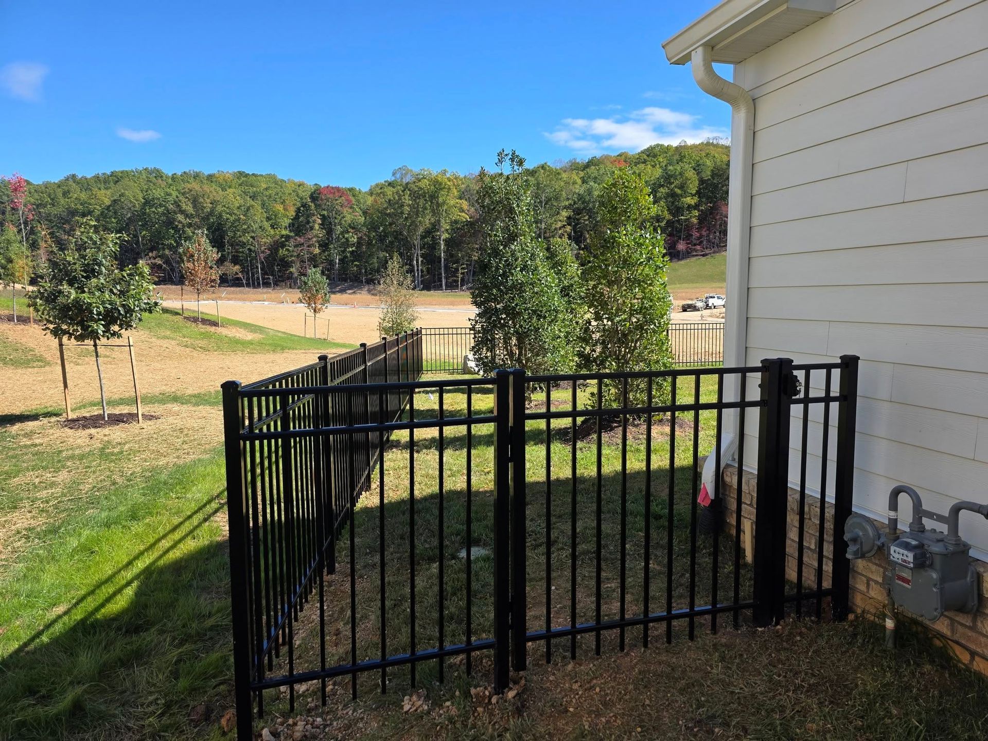 Black metal fence surrounding a yard with grass, near a house with white siding and a wooded area.