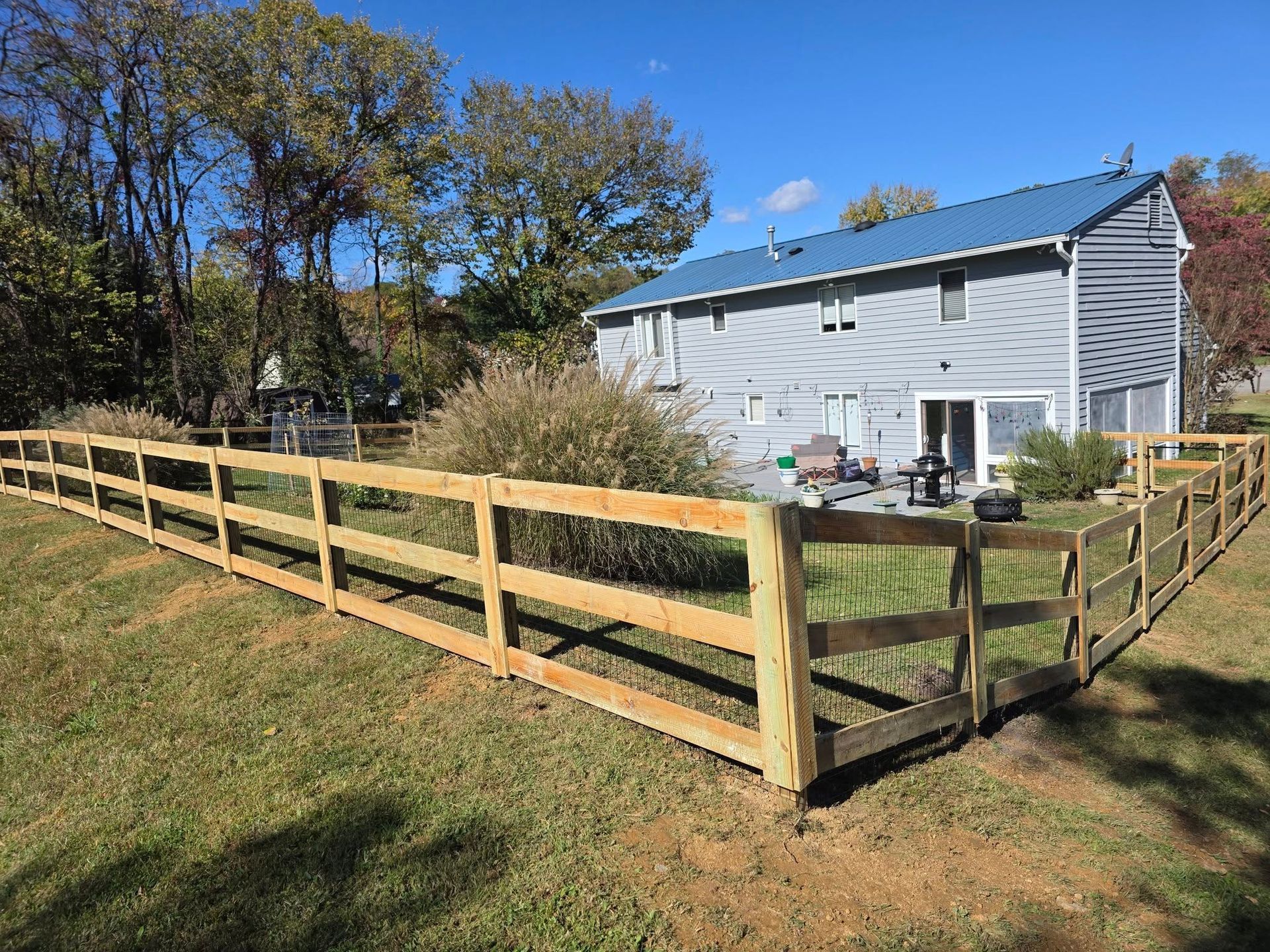 A wooden split-rail fence surrounds a grassy yard, with a two-story gray house in the background under a blue sky.