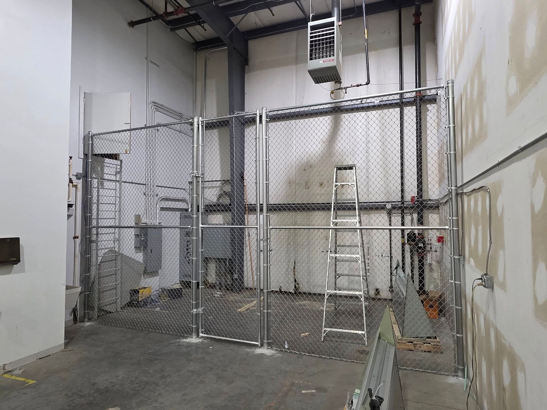 Chain-link fence enclosure in a building's interior. A ladder leans against the fence near electrical panels.
