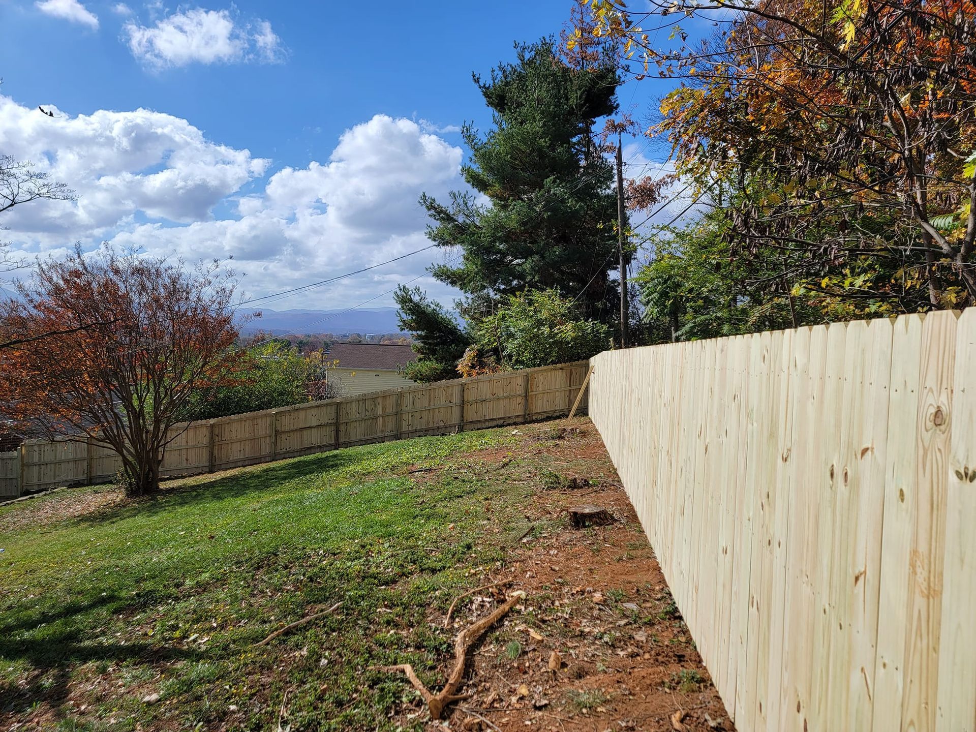 Wooden fence along a grassy yard with autumn trees and a partly cloudy blue sky.
