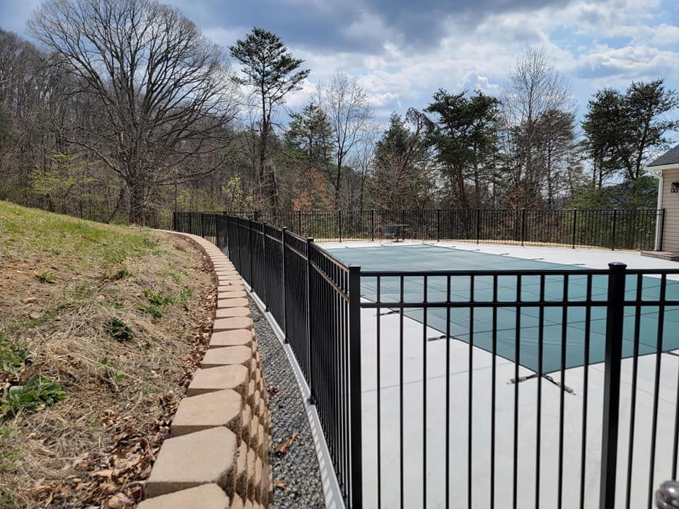 Black metal fence surrounds a pool covered with a blue tarp, on a hillside with trees and cloudy sky.