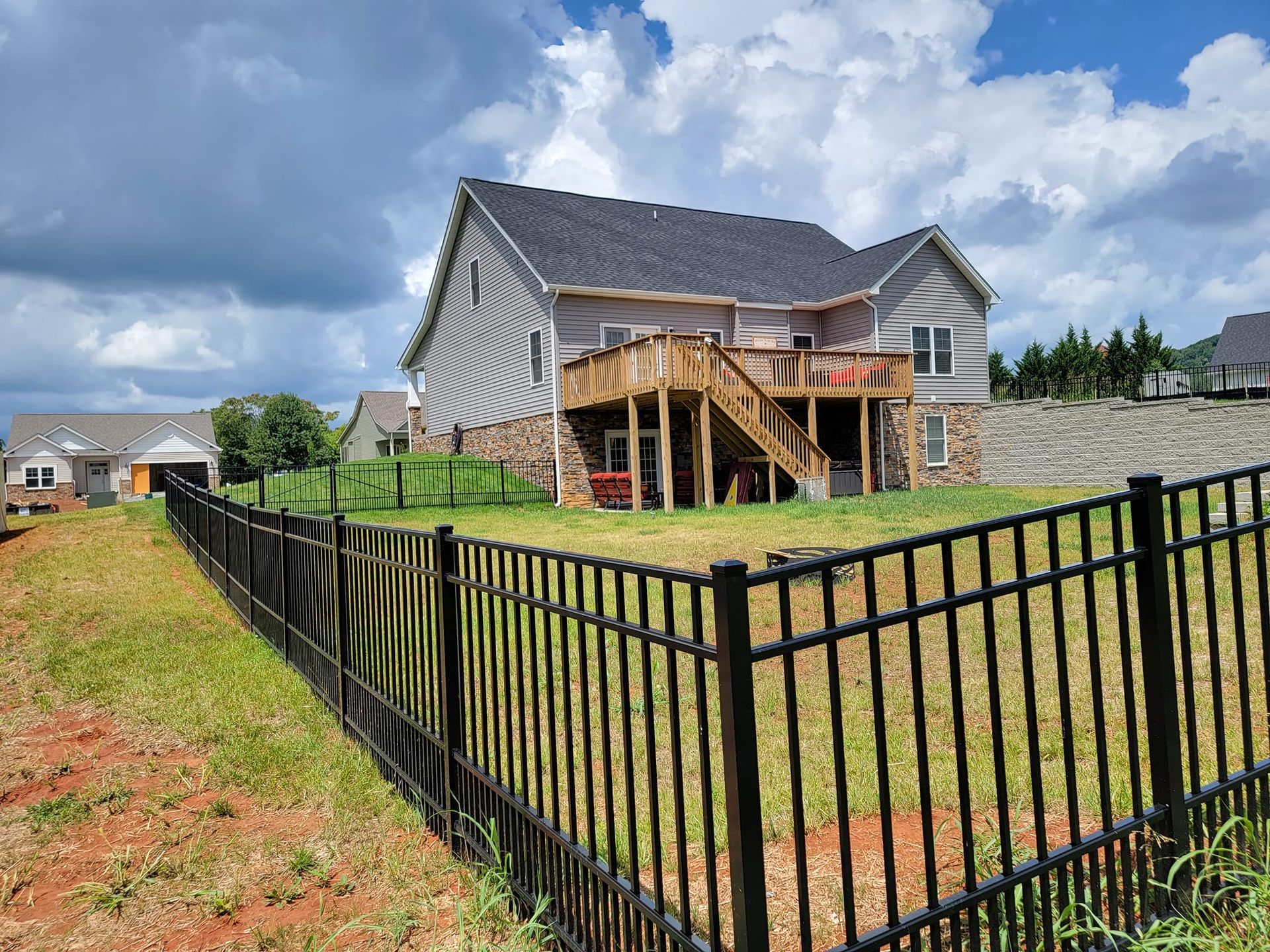 Backyard of a two-story house with a wooden deck, black fence, and cloudy sky.