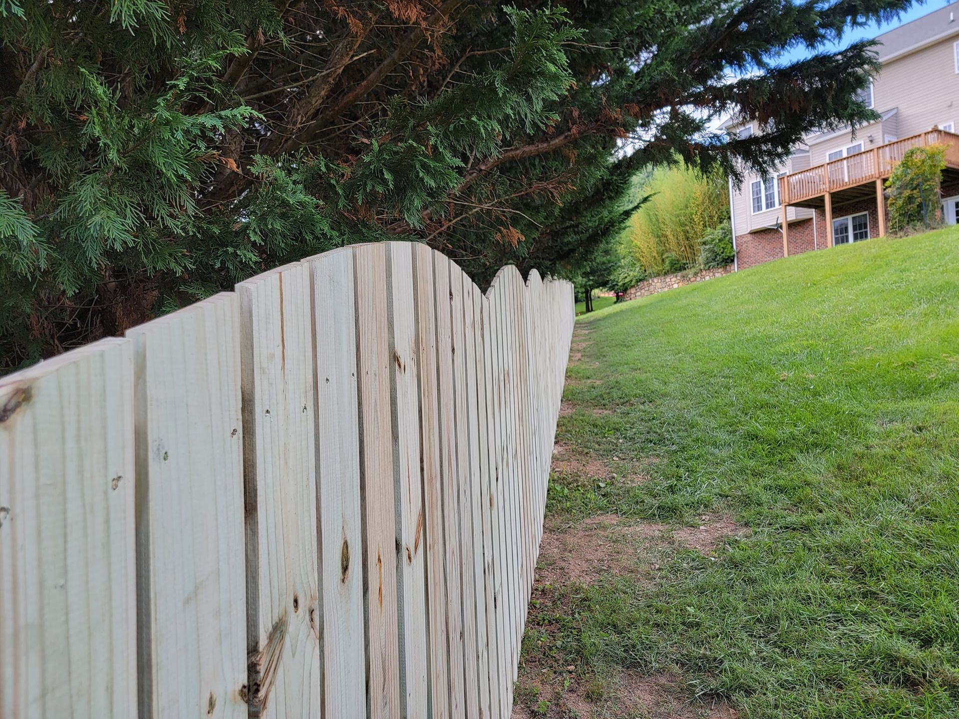 Wooden fence curving along a grassy hill, with a house and trees in the background.