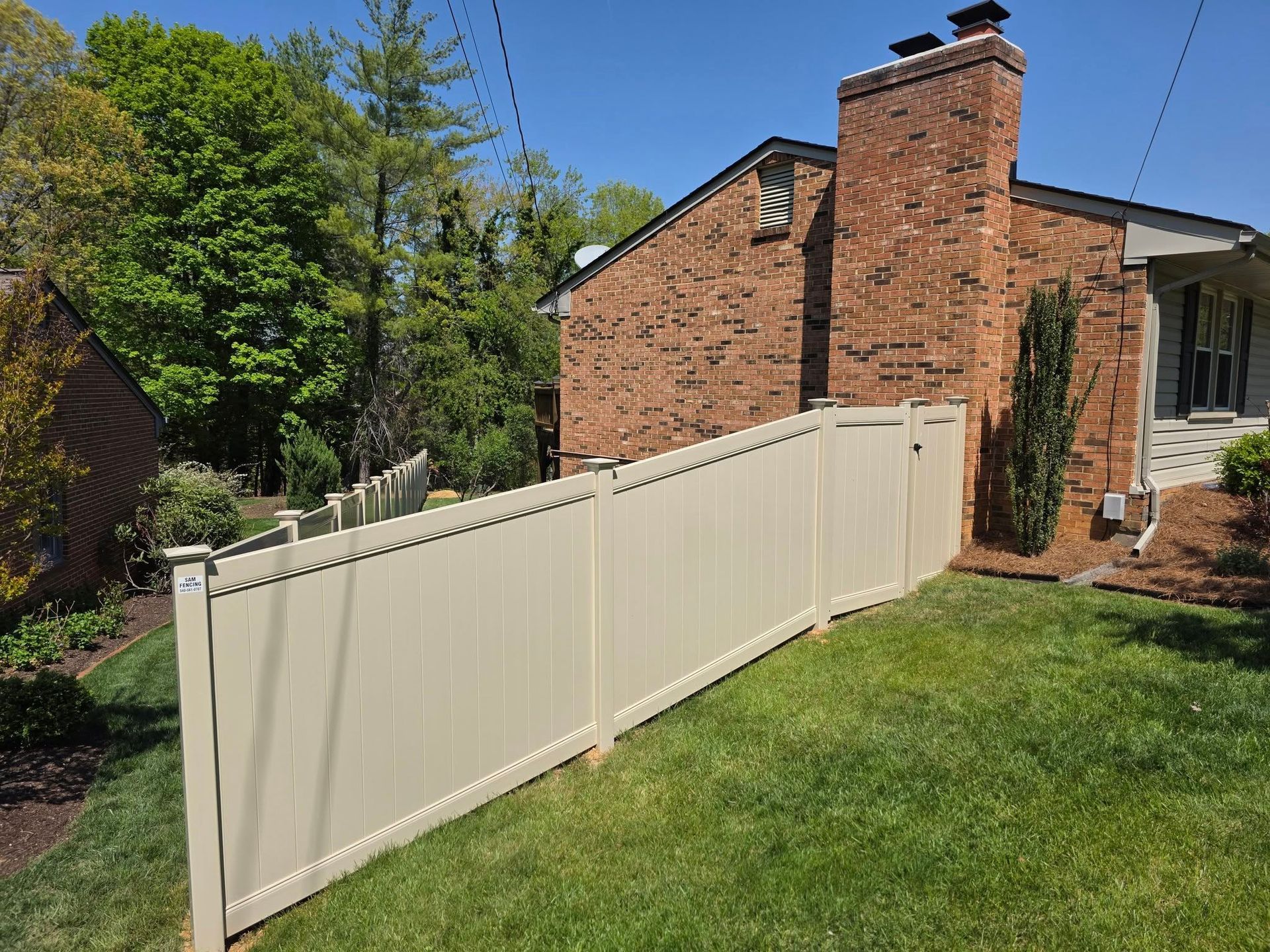 Cream-colored fence curving around a grassy yard next to a brick house and trees under a clear blue sky.