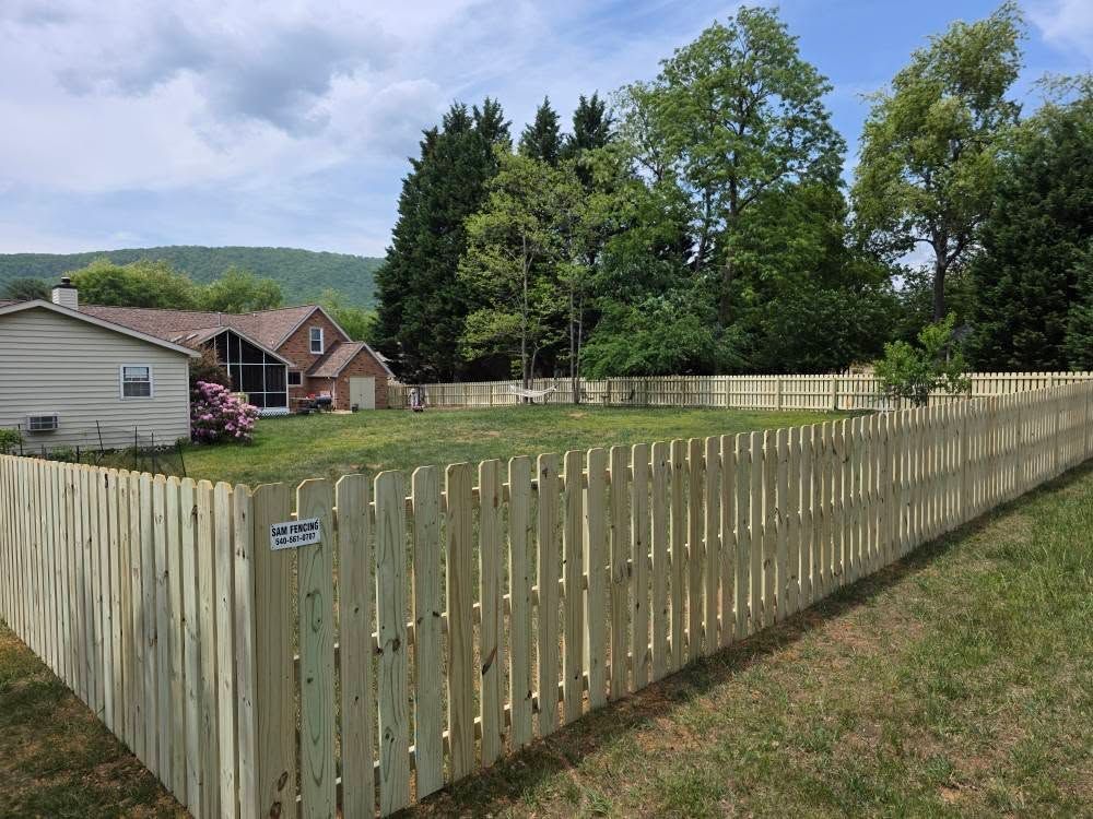 Wooden picket fence around a grassy backyard with two houses and trees under a blue sky.