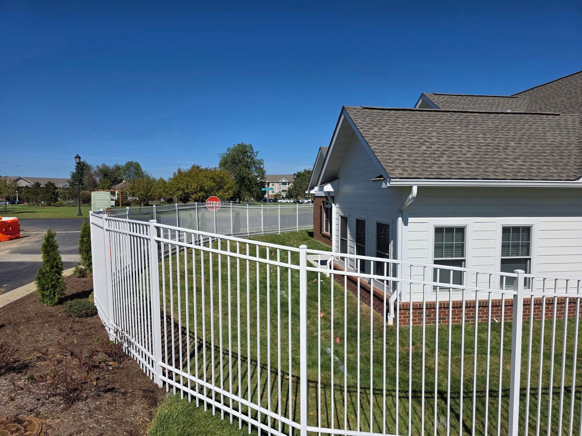 White fence curving in front of a white building with brown roof, green grass and blue sky.