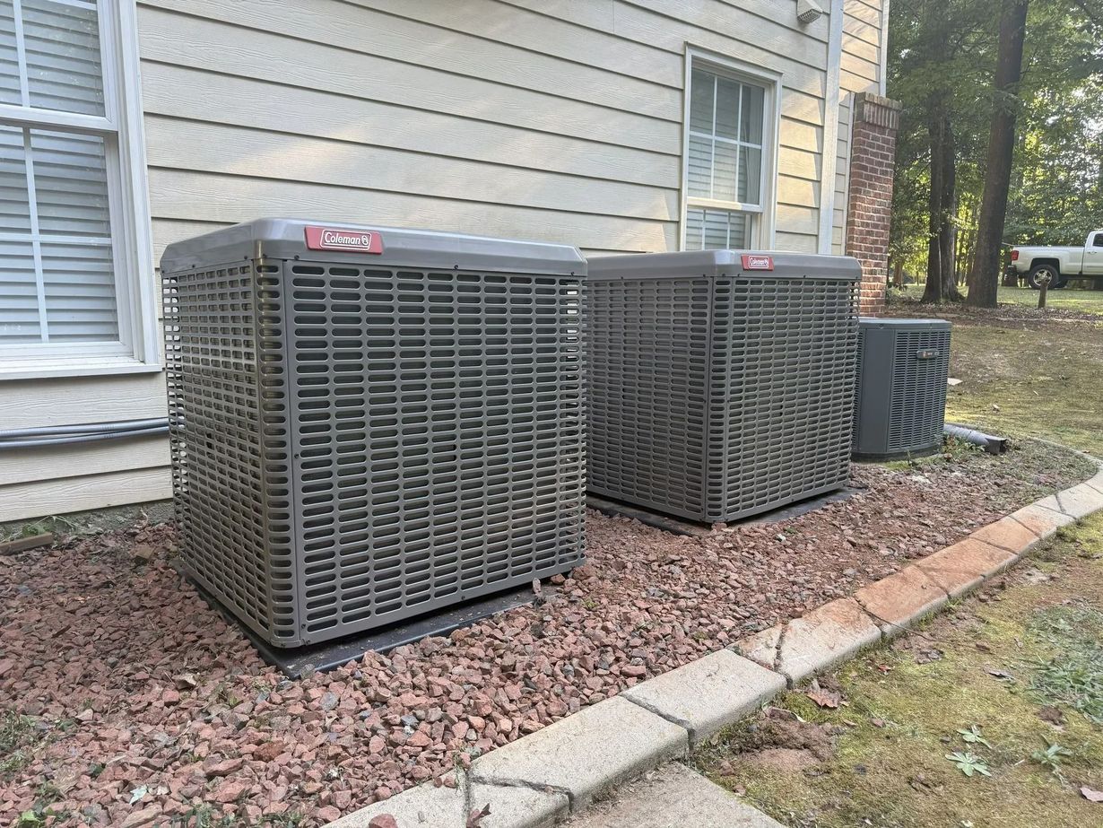 Two gray HVAC units sit on a bed of brown stones next to a house with a window and a brick chimney.