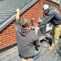 Two workers on a roof by equipment; one in gray hoodie, the other in a black cap. Brick building.