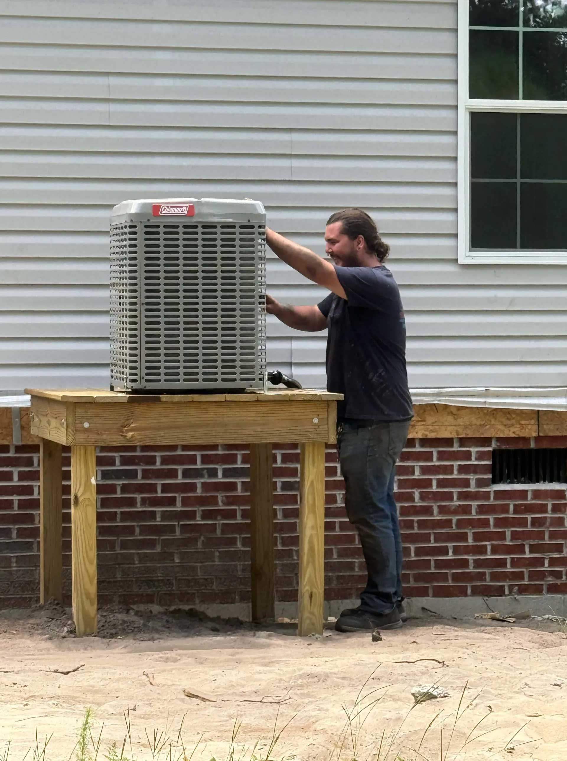 Man installing an air conditioner unit on a wooden table next to a brick foundation and siding.