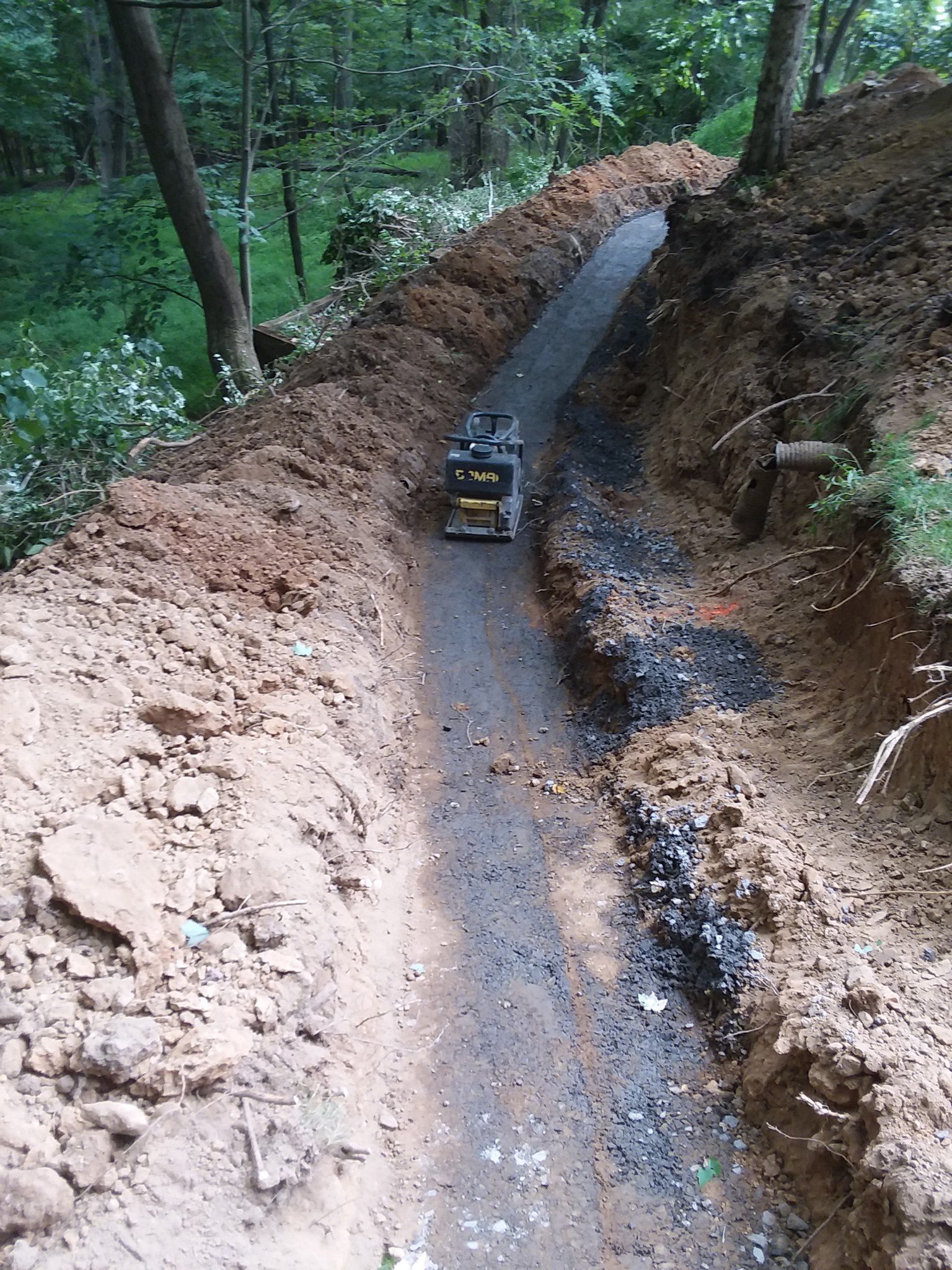 Narrow gravel path under construction through a wooded area; compactor machine in use.