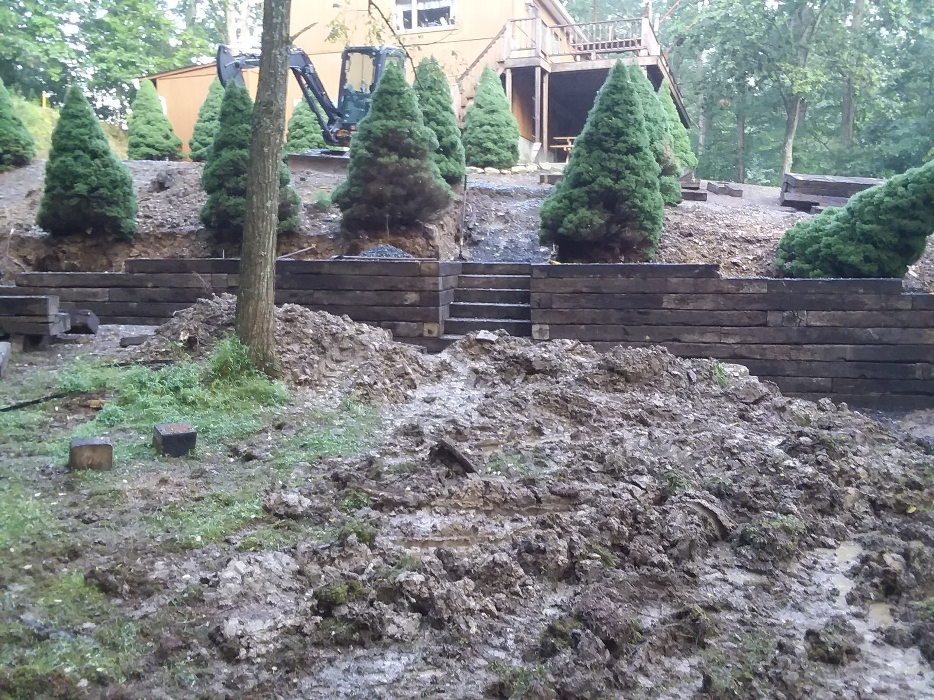 Muddy construction site with retaining wall, stairs, evergreen trees, and heavy machinery.