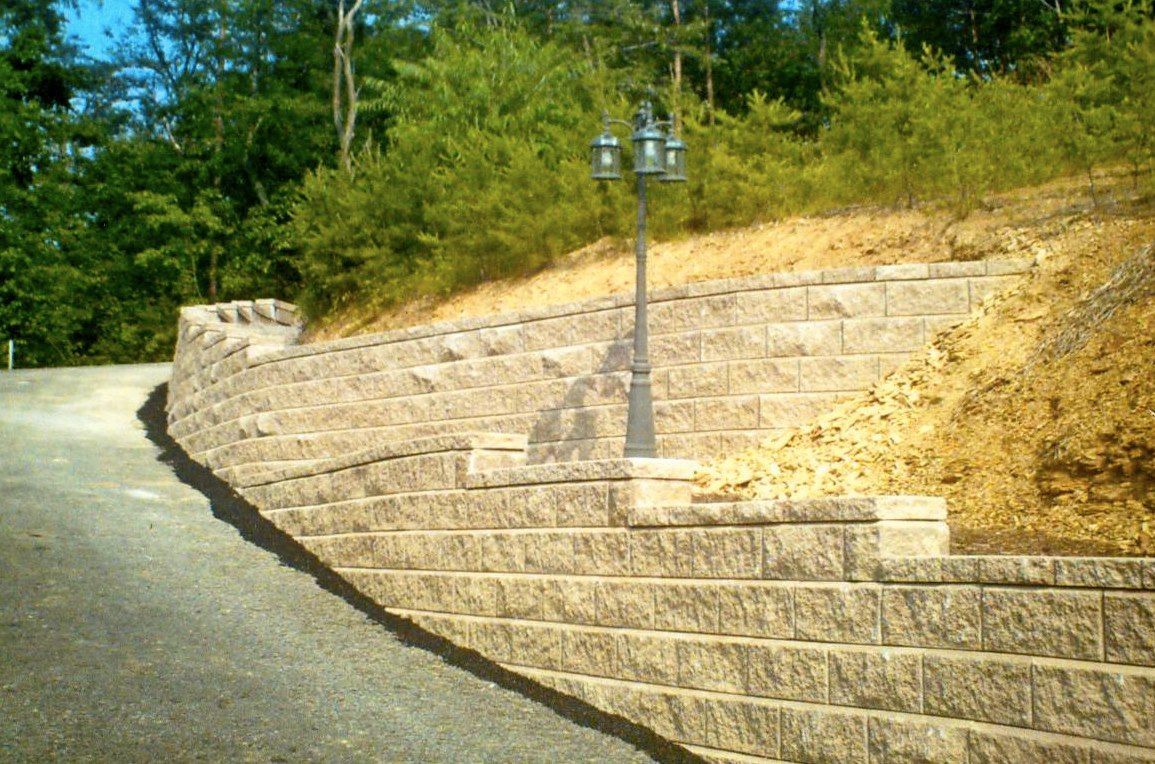 Stone retaining wall along a road. A lamp post is mounted in the wall. Green trees in the background.