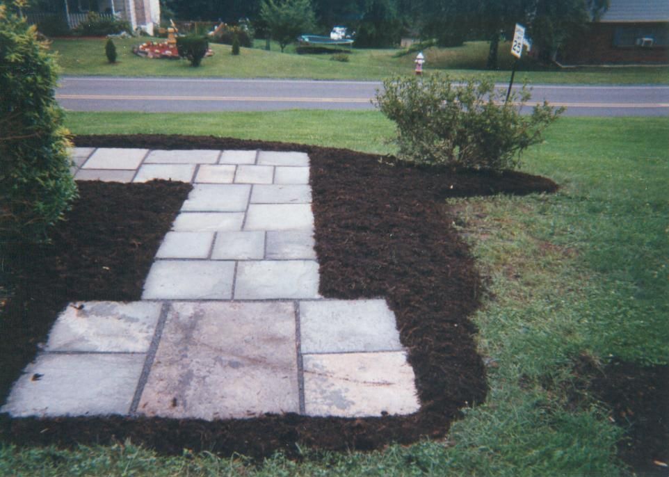 Stone walkway lined with dark mulch in a grassy yard, with a road and bushes in the background.