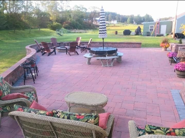 Red brick patio with wicker furniture, a fire pit, and an umbrella, overlooking a green lawn.