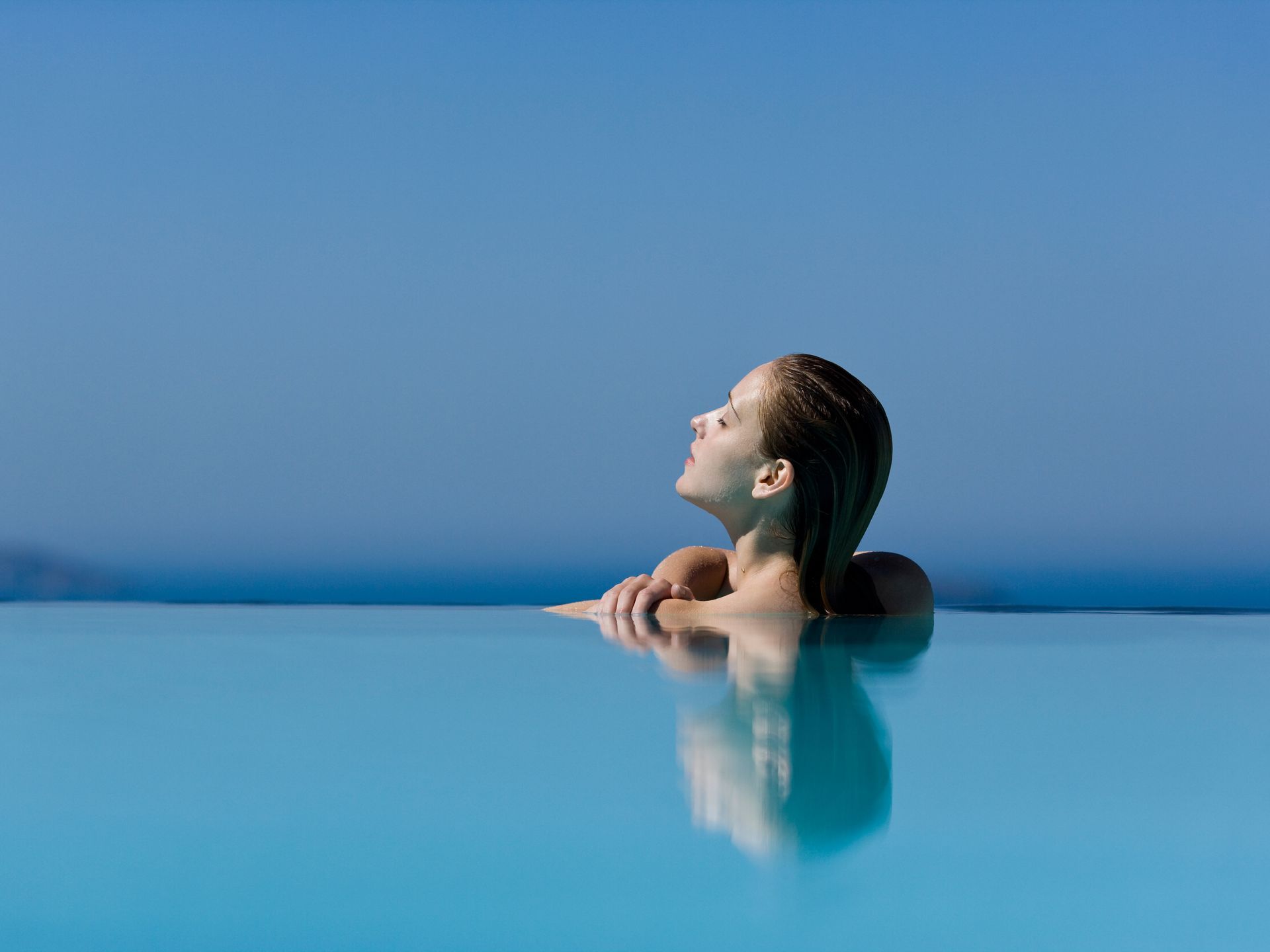 Person relaxing in an infinity pool at sunset, looking upward over calm blue water.