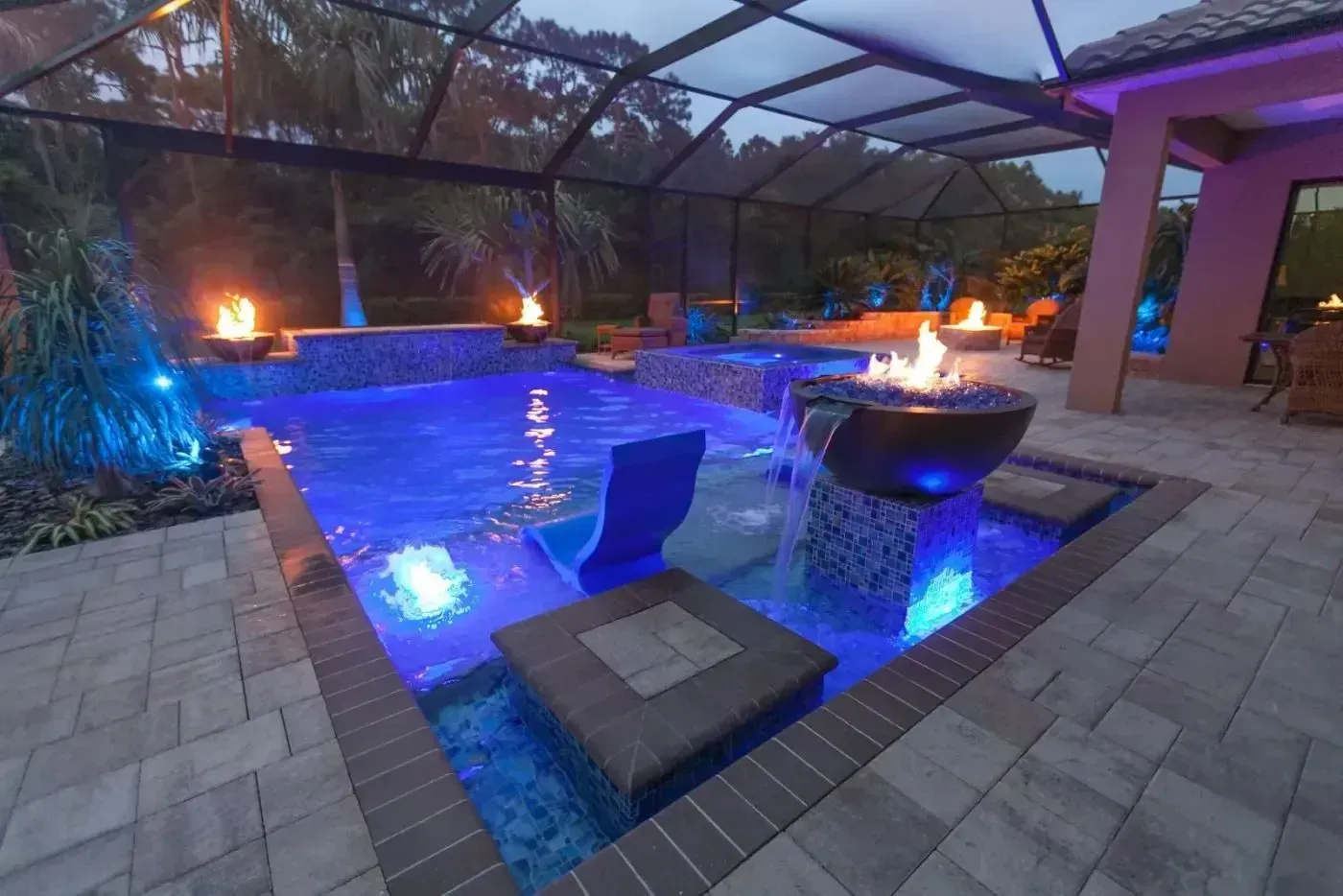 Illuminated backyard pool with blue water, fire bowl, and covered patio at dusk