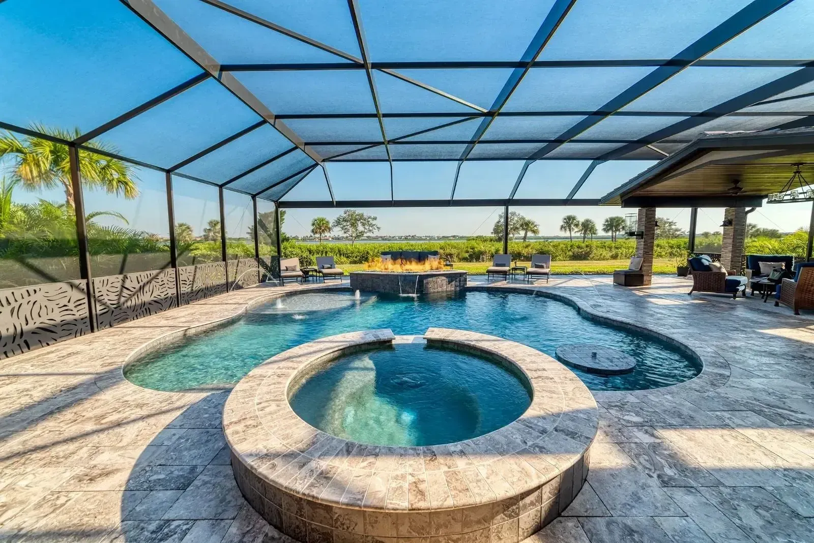 Covered outdoor pool with spa, blue water, stone deck, and screened patio overlooking a sunny landscape