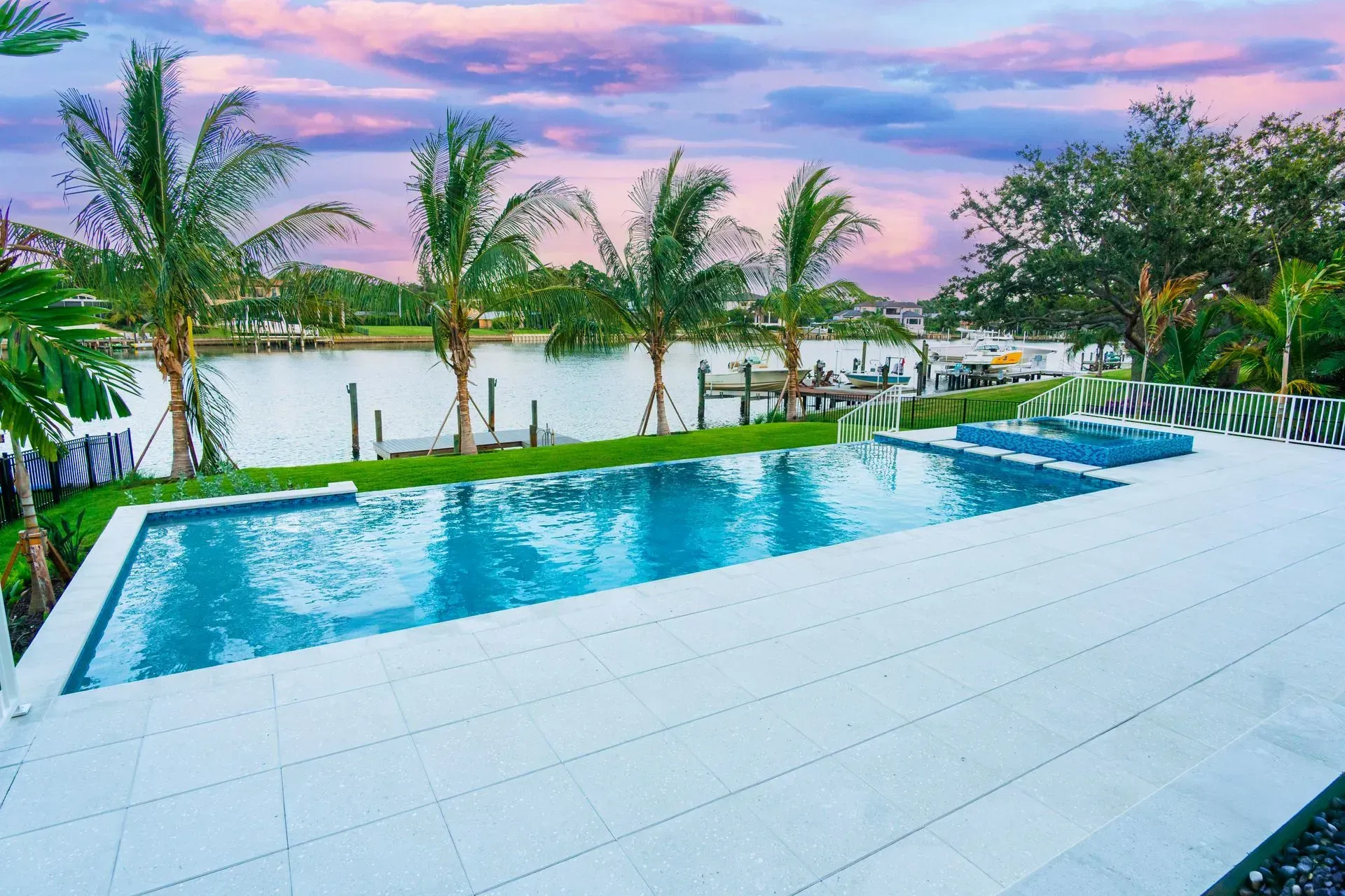 Infinity pool overlooking palm trees and calm water at sunset, with a pink and purple sky