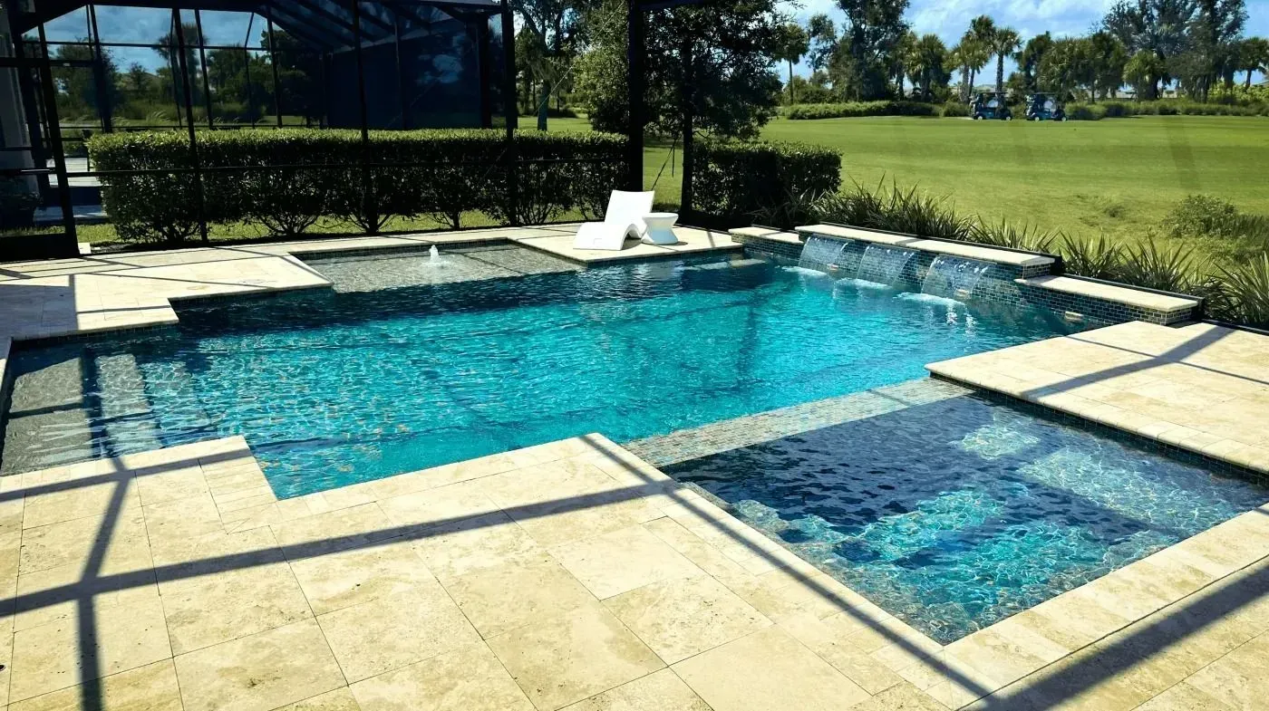 Backyard swimming pool with blue water, tan stone decking, and a grassy lawn beyond.