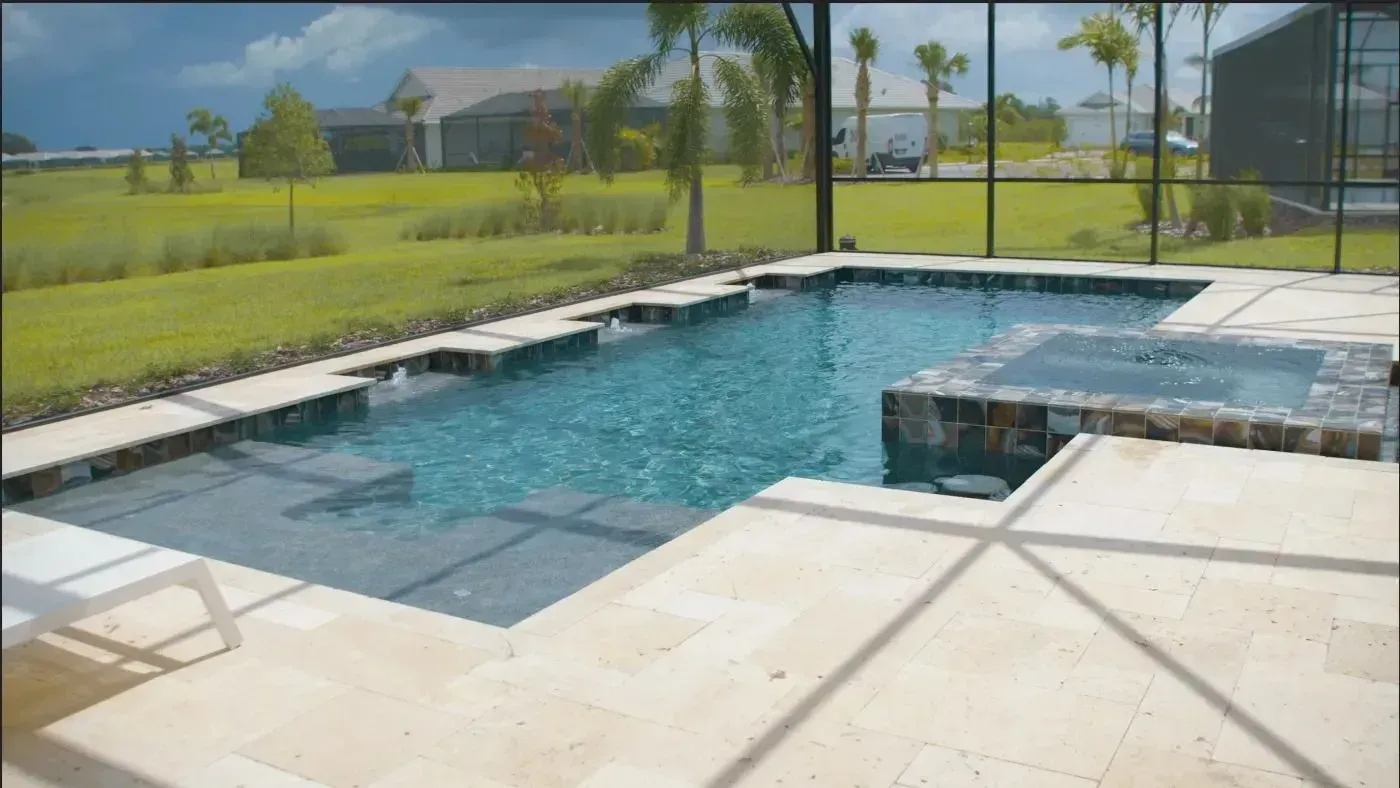 Backyard swimming pool with stone patio, glass fence, and grassy lawn under sunny blue sky