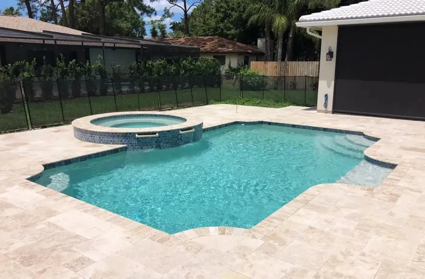 Backyard swimming pool with a raised hot tub and beige stone deck beside a garage