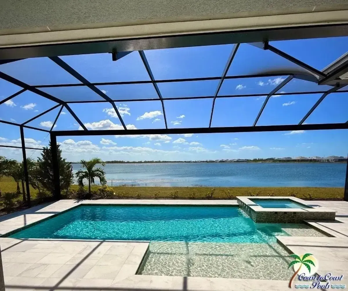 Covered patio with a turquoise pool and spa overlooking a calm lake under a blue sky