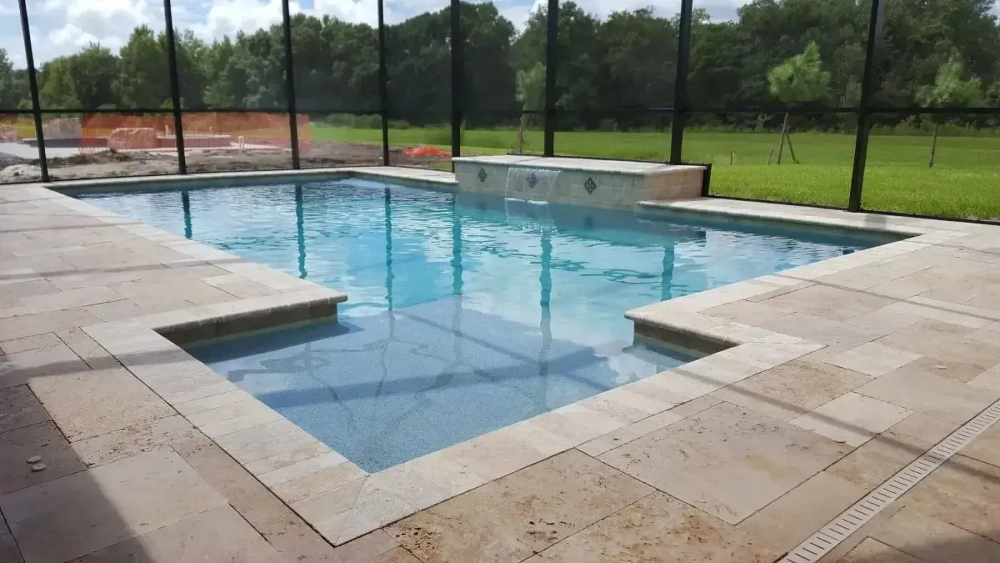 Outdoor swimming pool with clear blue water, beige stone deck, and black metal fence in a grassy park setting