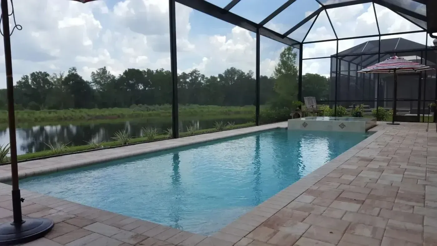 Covered outdoor pool beside a brick deck, with pond and trees beyond the screened enclosure.