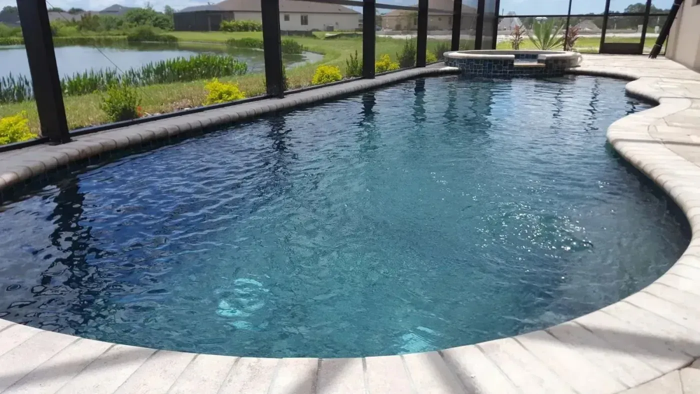 Curved backyard pool with clear blue water, white stone deck, and lake view through screened enclosure