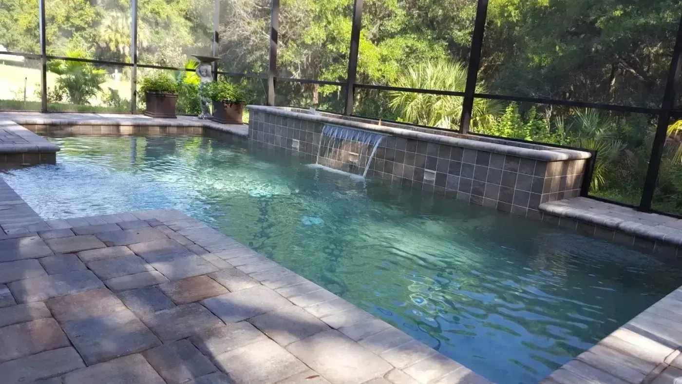 Rectangular outdoor pool with stone deck and black safety fence, surrounded by trees and greenery