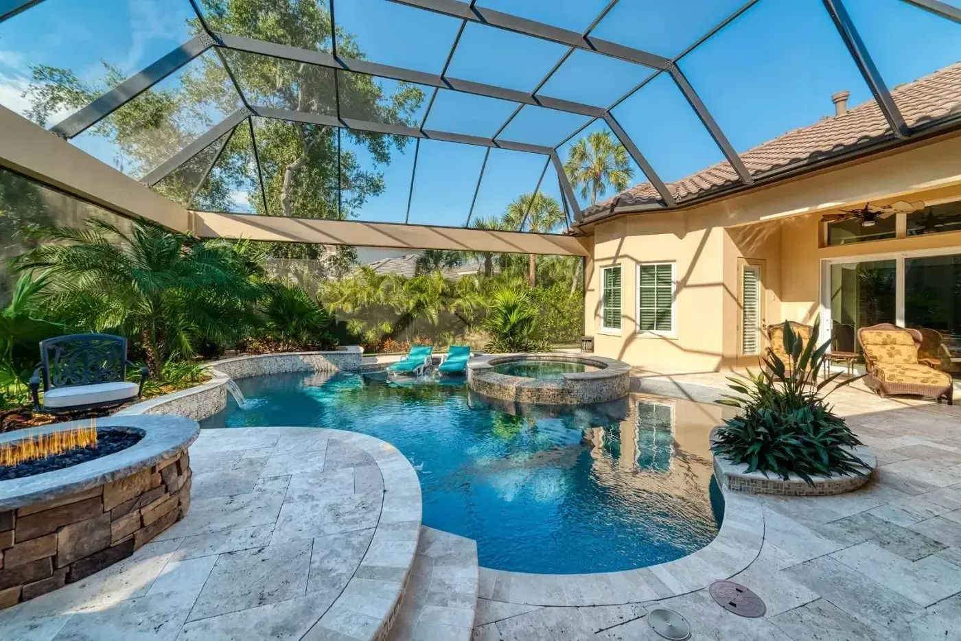 Screened backyard pool with stone patio, tropical landscaping, and a beige house under blue sky.