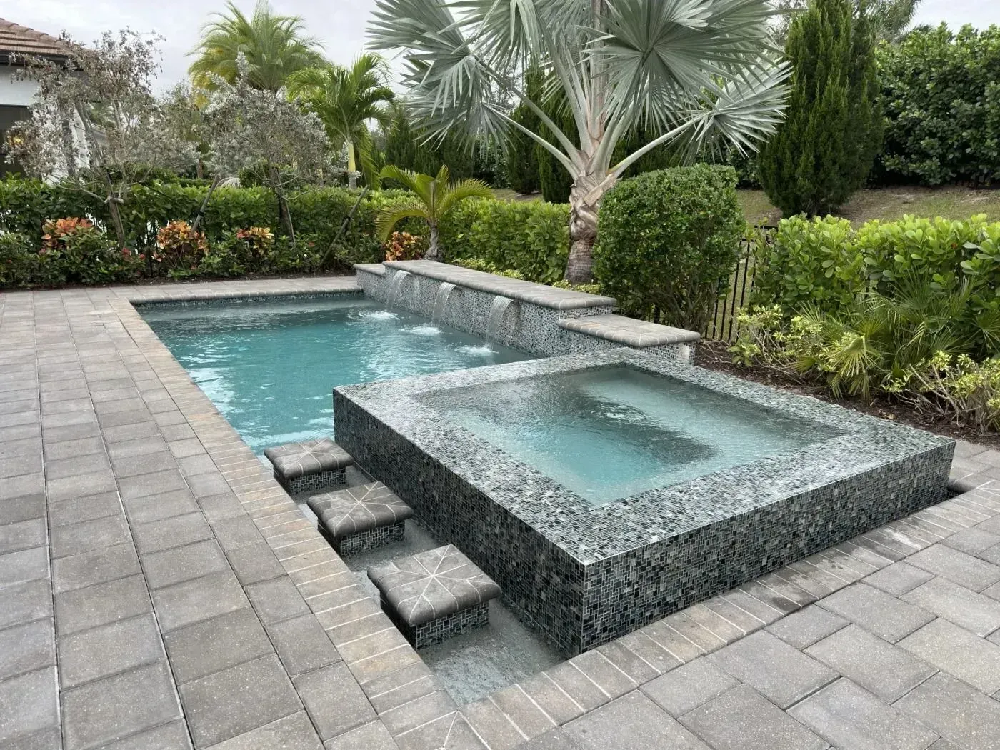 Backyard pool with a raised square spa, stone pavers, and lush tropical landscaping