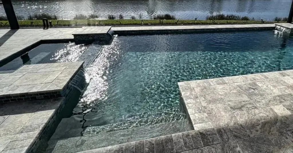 Modern backyard swimming pool with a shallow shelf, stone deck, and water features beside a lake.