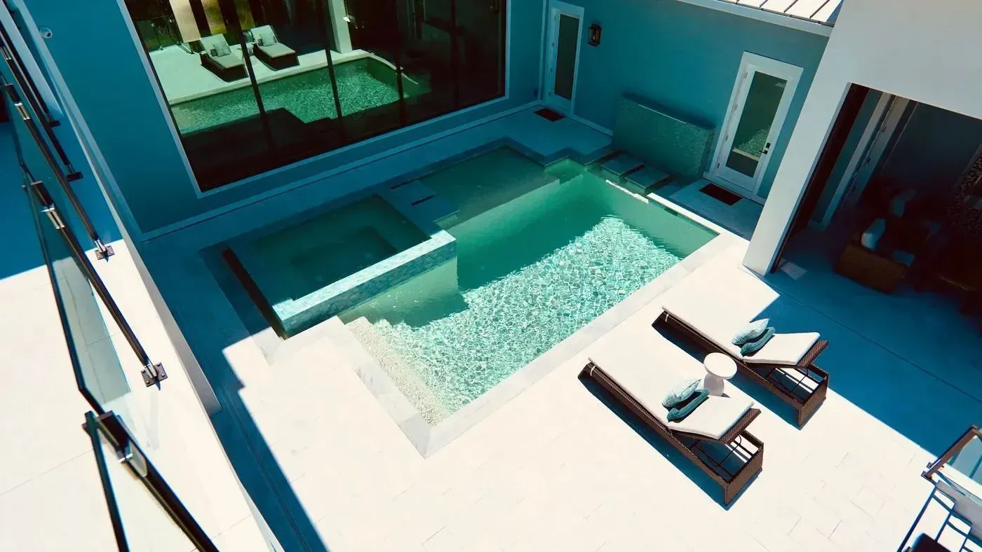 Modern patio with a small turquoise pool, white lounge chairs, and blue walls in bright sunlight