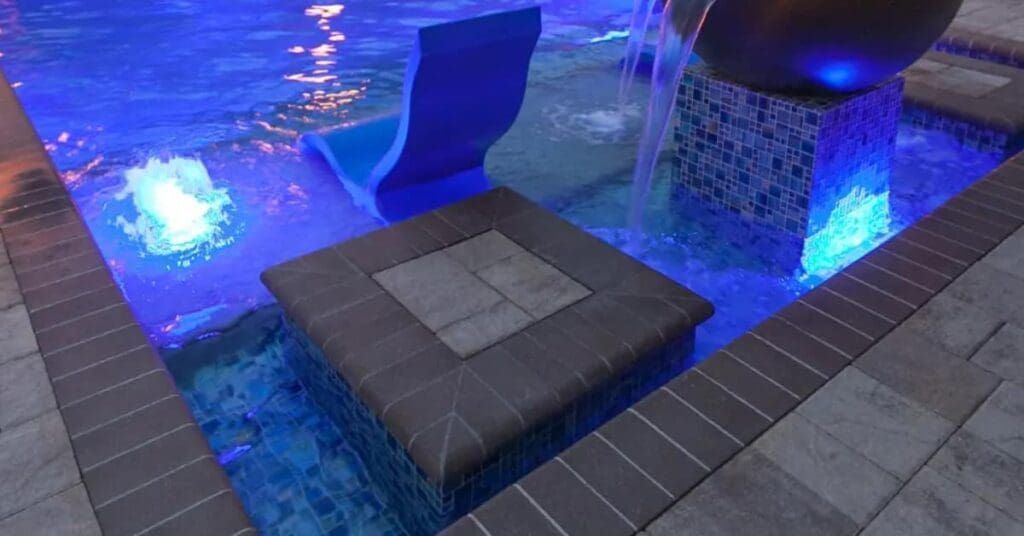 Blue-lit water feature with stone platforms and fountains beside a tiled walkway