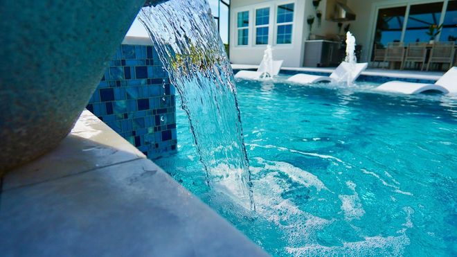 Water flowing from a tiled pool spout into a bright blue swimming pool with fountains and a house in the background