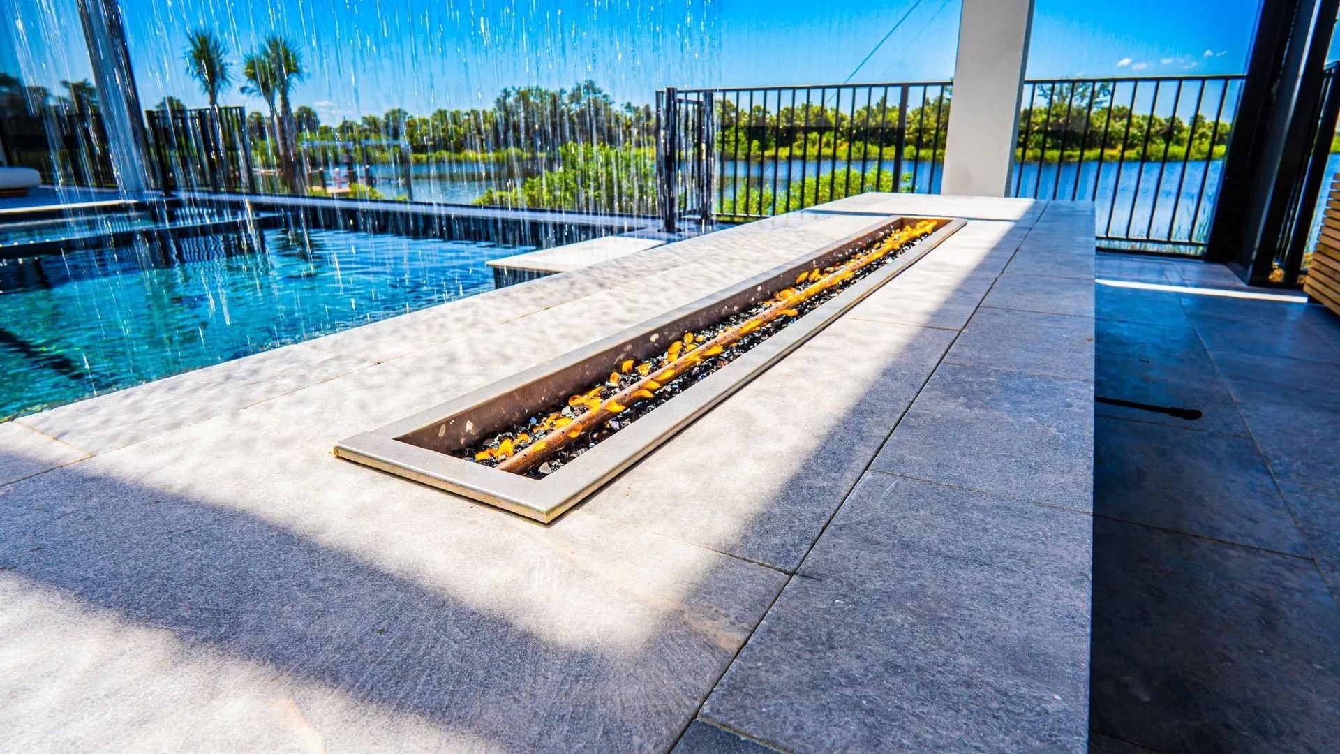 Modern outdoor fire pit on a tiled patio beside a pool and black metal fence, with blue sky and trees beyond.