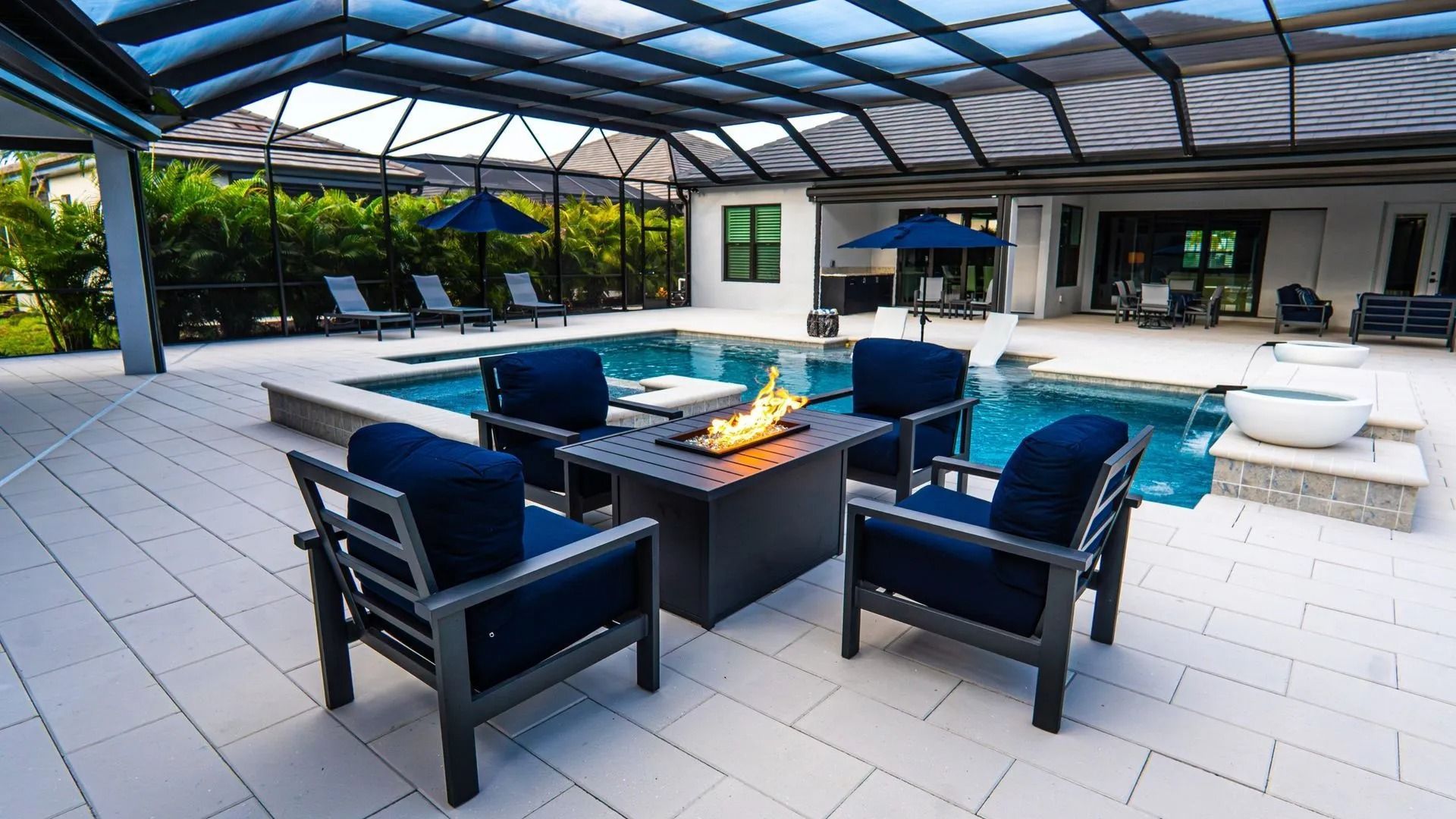 Modern patio with black chairs around a fire pit under a glass roof, beside a pool and white loungers