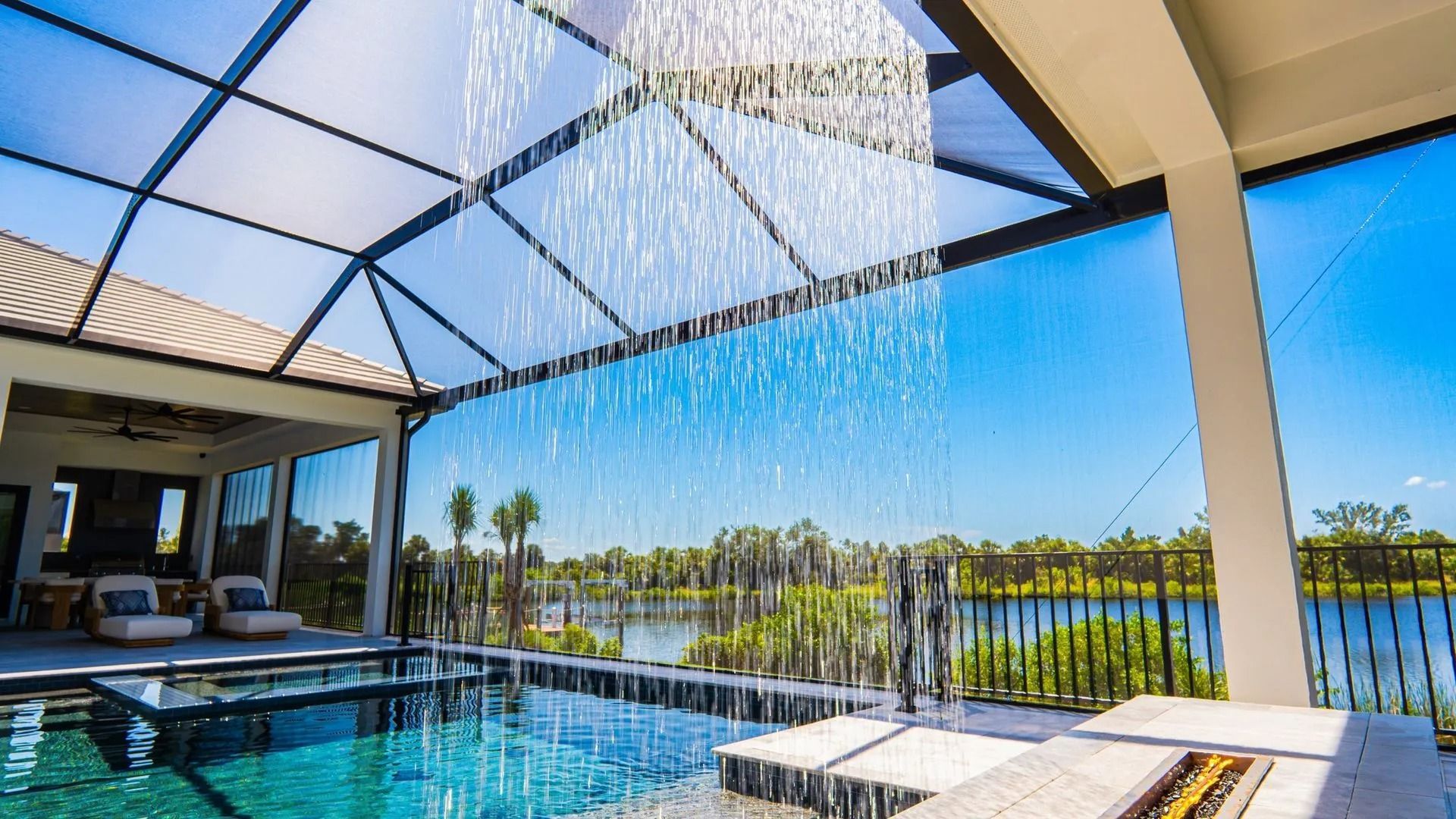 Poolside patio with a covered waterfall feature, lounge chairs, and a view of water and palm trees