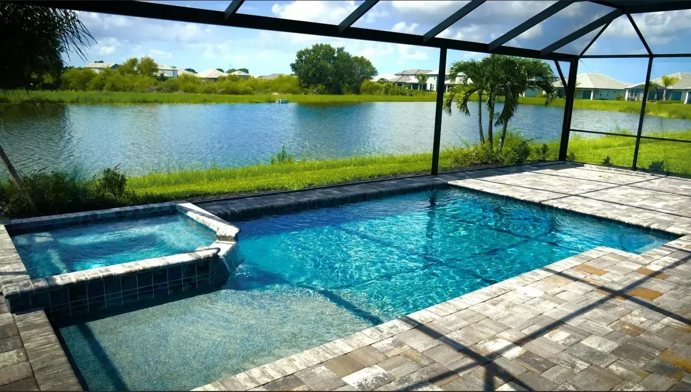 Covered backyard pool beside a lake, with blue water, stone patio, and screened enclosure