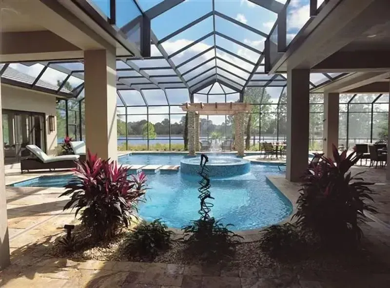 Indoor pool under a glass roof, with lounge chairs and tropical plants overlooking a lake outdoors