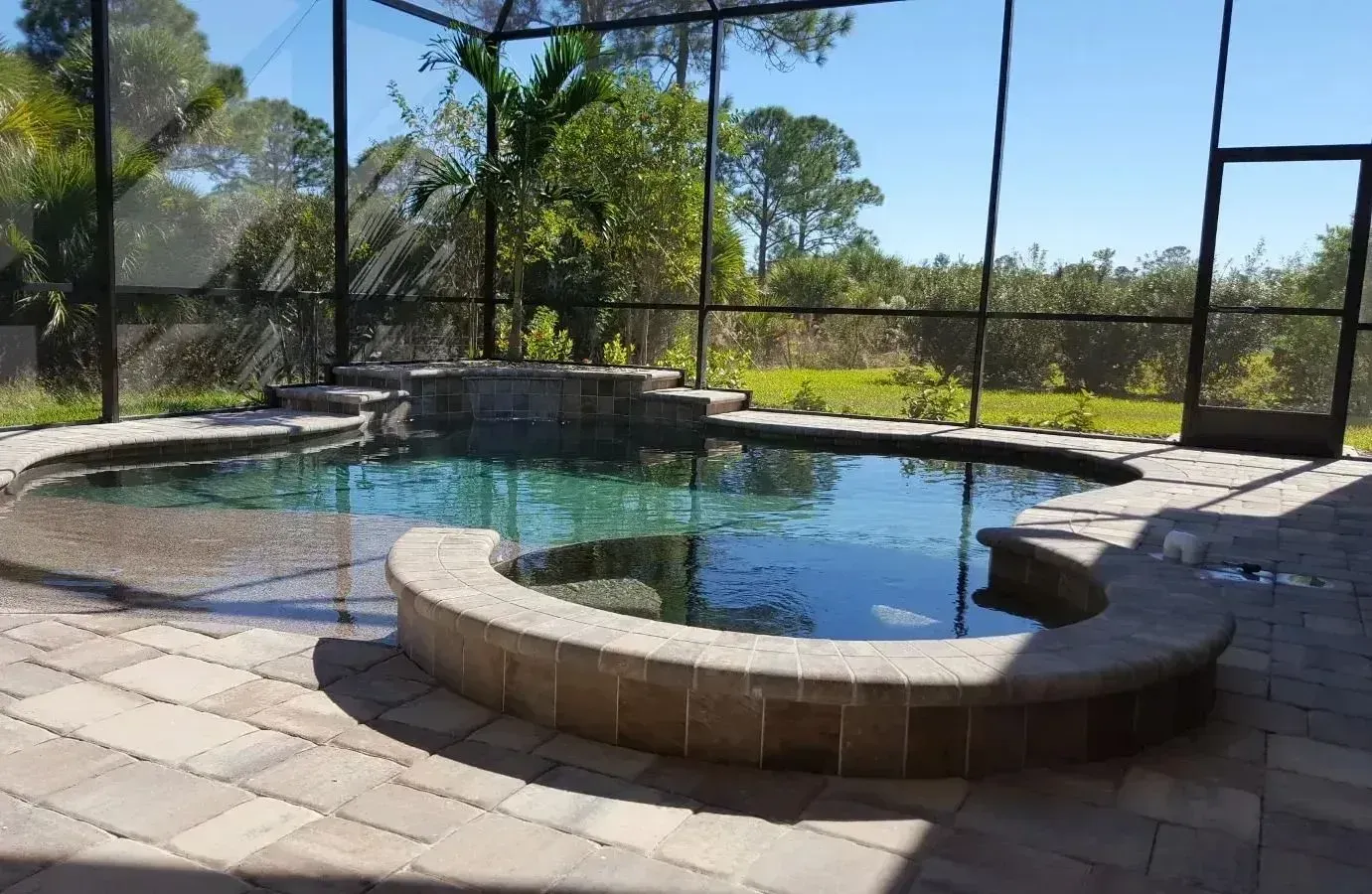 Screened patio with a curved stone pool and spa, overlooking trees and blue sky