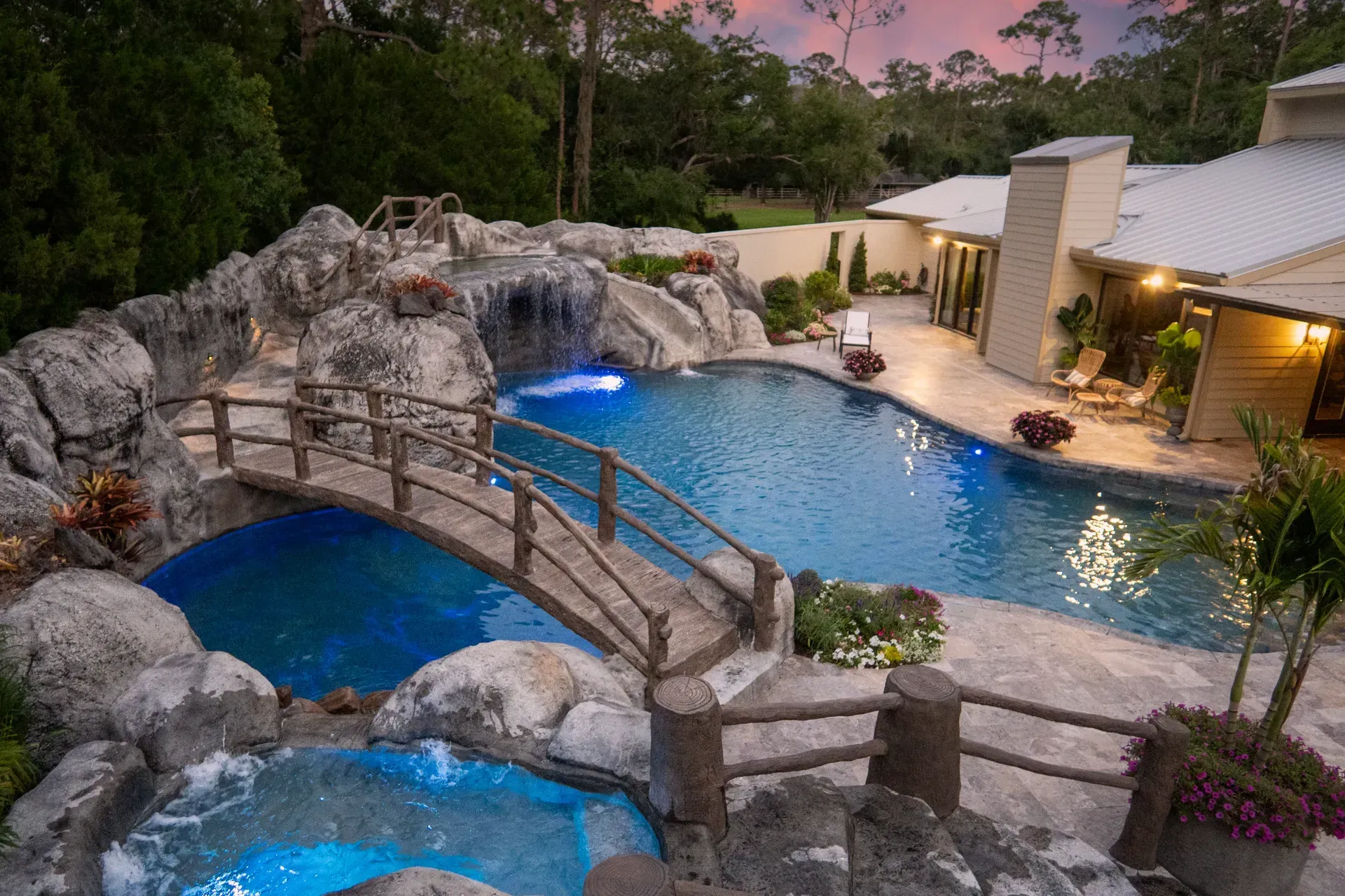 Luxury backyard pool with rock waterfall, blue water, and wooden bridges at dusk