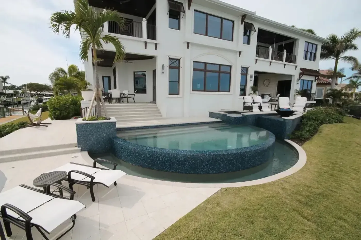 Modern white house with a curved blue-tiled pool and lounge chairs on a patio, overlooking a grassy yard