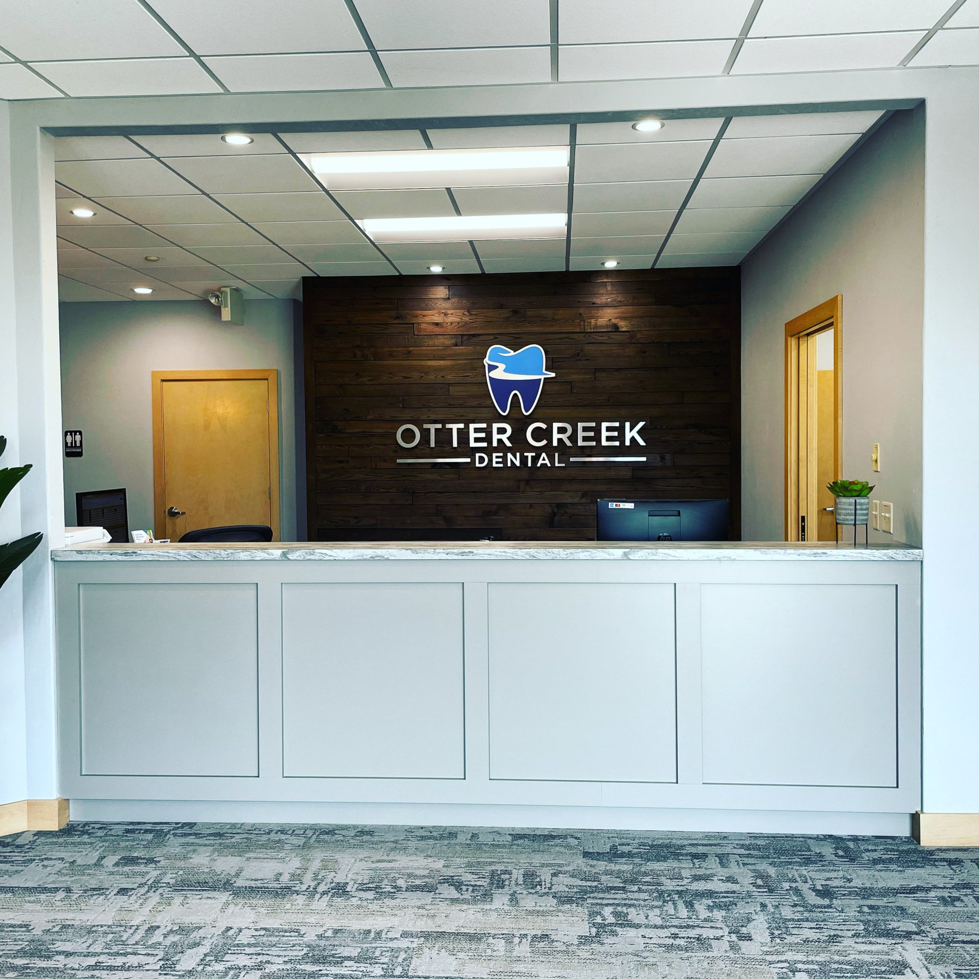 Reception area of Otter Creek Dental with a logo on a wood-paneled wall, gray counter, and gray flooring.
