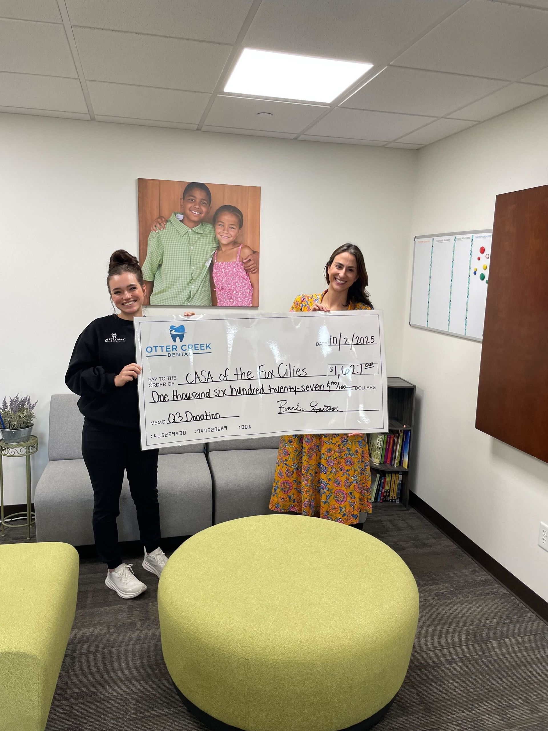 Two women holding an oversized check indoors. A photo of children hangs on the wall behind them.