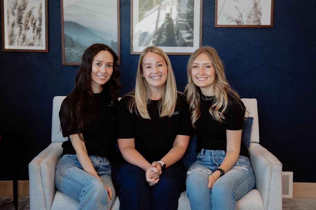 Three women in black tops and jeans sitting on a sofa in front of a blue wall with framed art.