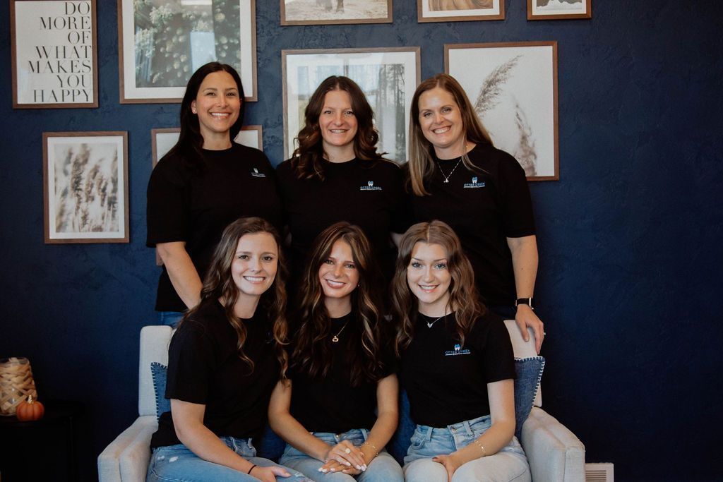 Group of six people smiling in front of a blue wall with framed artwork.