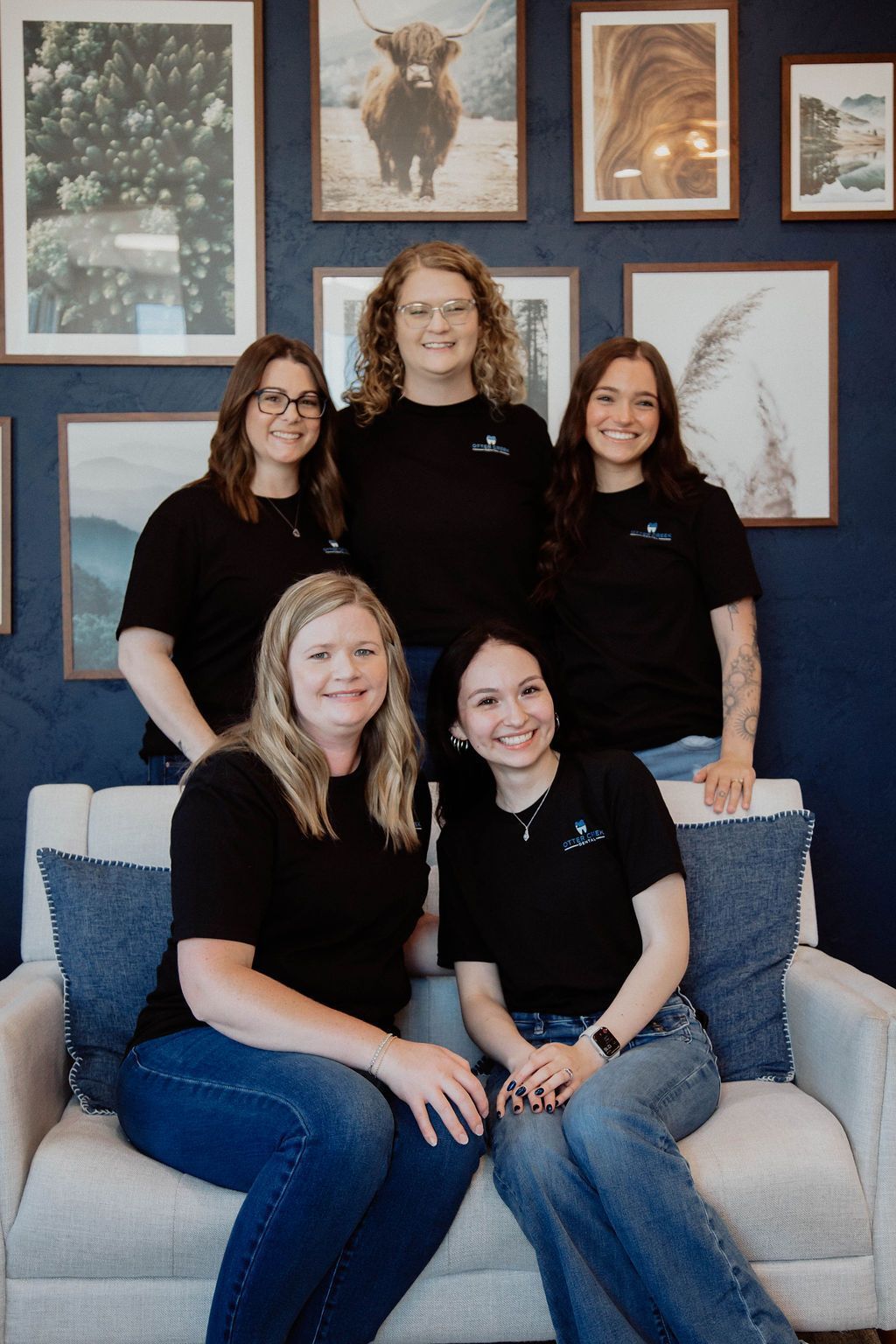 Five people in black t-shirts and jeans posing on a white couch in front of framed artwork.
