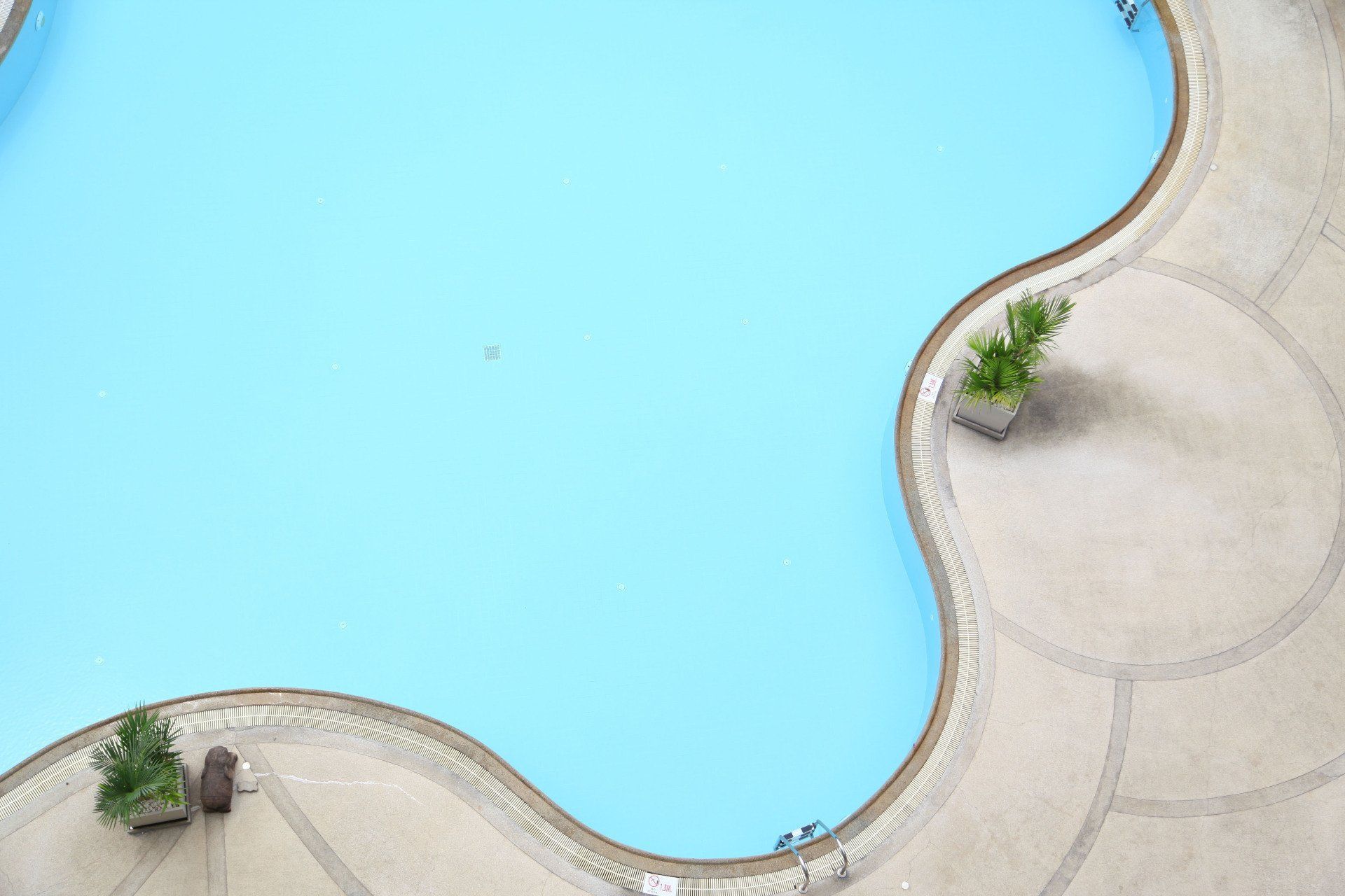A turquoise, kidney-shaped swimming pool surrounded by beige stone and a few potted plants.